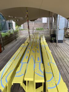 Photograph of large bright yellow painted picnic benches on a rooftop terrace. The yellow benches have been painted with a sky-blue illustration of a person with long arms and legs, in a kitsch and fun style. The floor is wooden decking, and around the edges there are large wooden planters filled with various plants. 